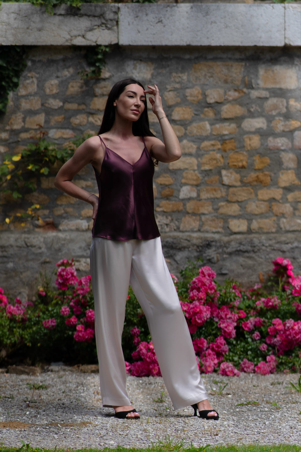 Woman in a purple top and white pants standing in front of a stone wall with pink flowers.